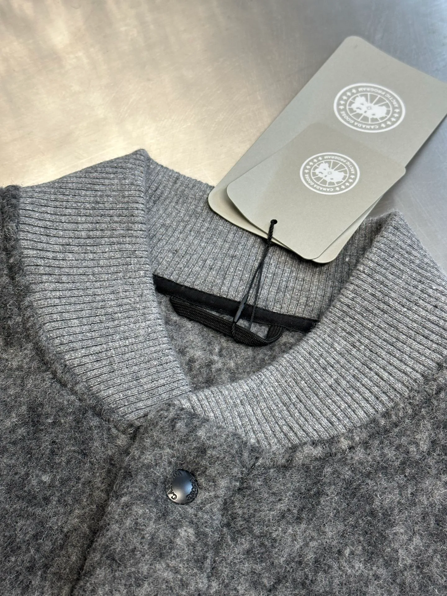P770 Canada Goose Fleece Badge Button Jacket The main fabric of this jacket is light gray wool blend fabric to improve the fabric's wear resistance, wrinkle resistance and warmth. Wool provides good warmth and a natural feel, while blended fibers enhance the fabric's practicality and durability.  The cutting process needs to ensure that the texture and color of the fabric are evenly distributed, especially with this light gray tone, to avoid chromatic aberrations. The stretchability of wool-blend fabrics needs to be taken into consideration when cutting pieces to ensure dimensional stability of the finished garment.  Use a sewing needle and thread suitable for wool-blend fabrics to avoid damage to the fabric during sewing. The sewing stitches are even and firm to prevent thread stripping.  The temperature needs to be controlled during ironing to avoid high temperature damage to the fabric while keeping the fabric smooth and crisp. The front and back body parts, sleeves and other main parts are stitched with flat seams to ensure a smooth silhouette. Align the fabric texture at the splices to keep the look neat.  The white circular armband on the left arm is an important part of the brand identity and is finely sewn to ensure it is strong and flat.  size：s m l xl